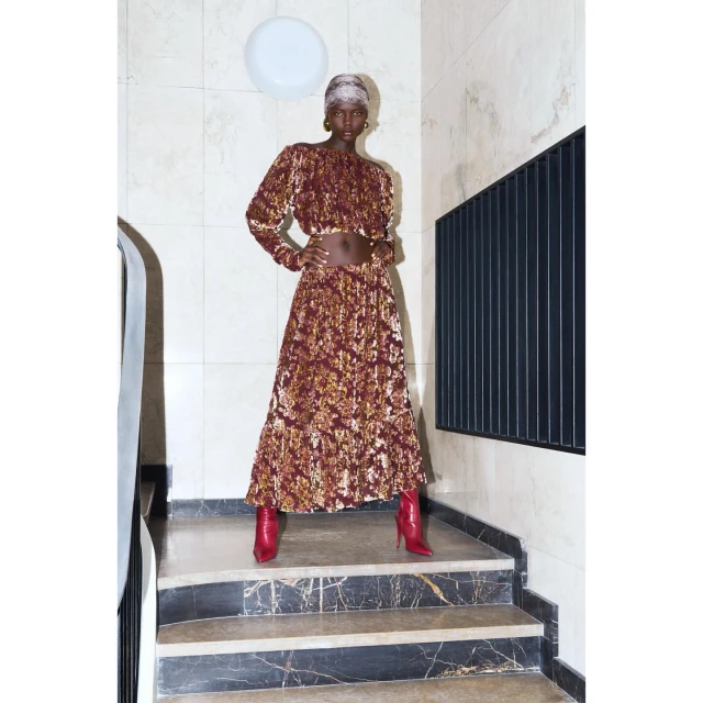 Full-length shot of a black female fashion model wearing a patterned blouse and skirt, posing on marble stairs in red boots.