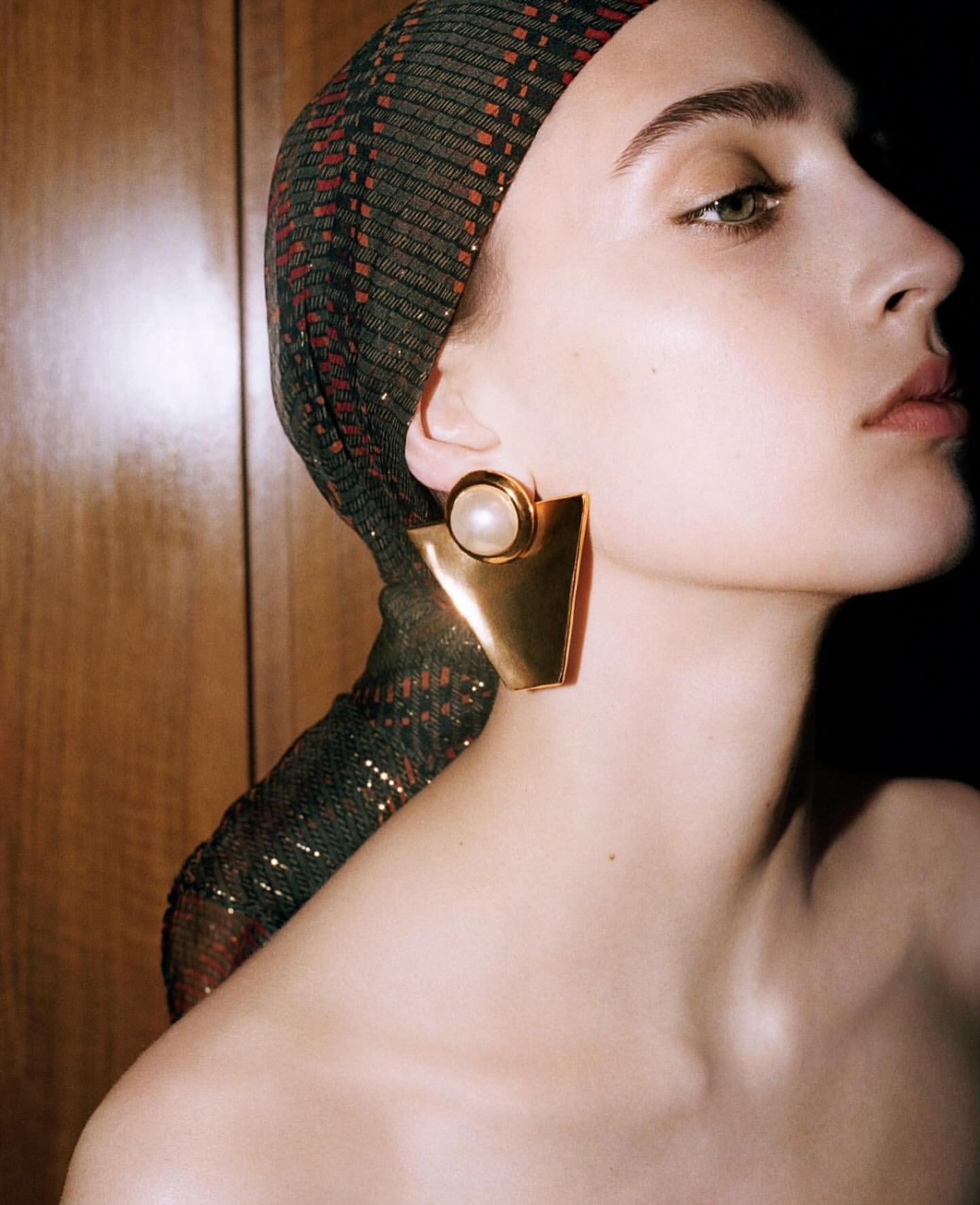 Adult woman wearing a sequin headscarf and gold statement earrings, shot in a photo studio with dark lighting and warm tones.