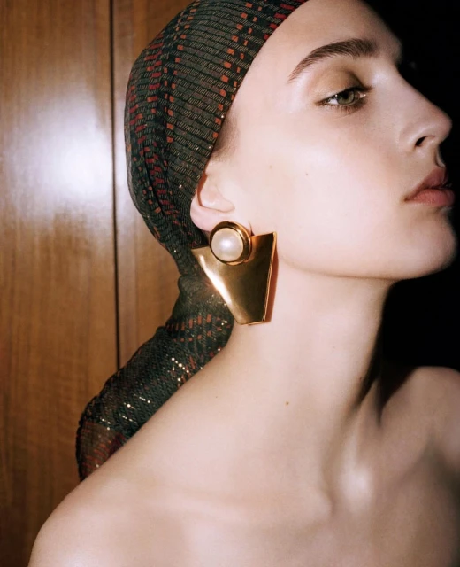 Adult woman wearing a sequin headscarf and gold statement earrings, shot in a photo studio with dark lighting and warm tones.