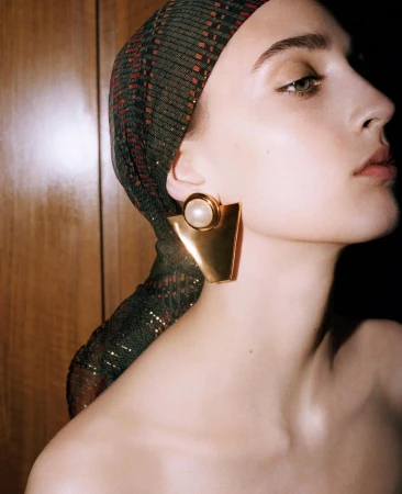 Adult woman wearing a sequin headscarf and gold statement earrings, shot in a photo studio with dark lighting and warm tones.