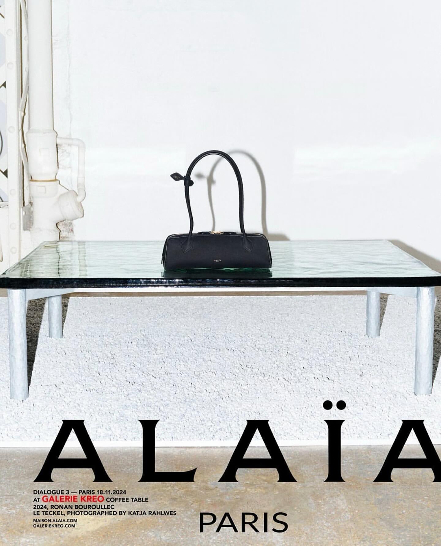 Alaïa handbag displayed on a glass table indoors, with natural lighting, against a textured white wall in a studio setting.
