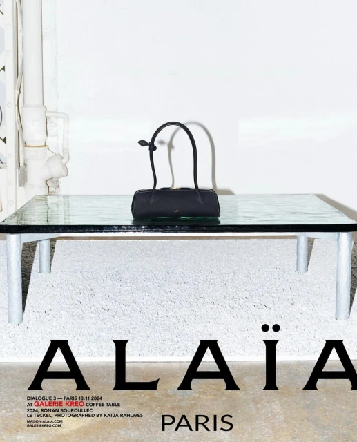 Alaïa handbag displayed on a glass table indoors, with natural lighting, against a textured white wall in a studio setting.