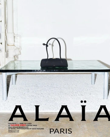 Alaïa handbag displayed on a glass table indoors, with natural lighting, against a textured white wall in a studio setting.