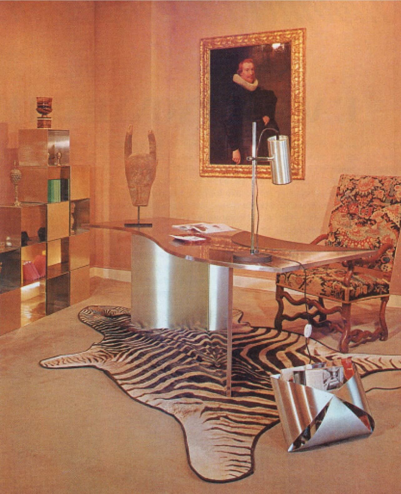 Interior of room featuring a desk with a lamp, ornate chair, zebra-print rug, reflective furniture, and a framed portrait on the wall.