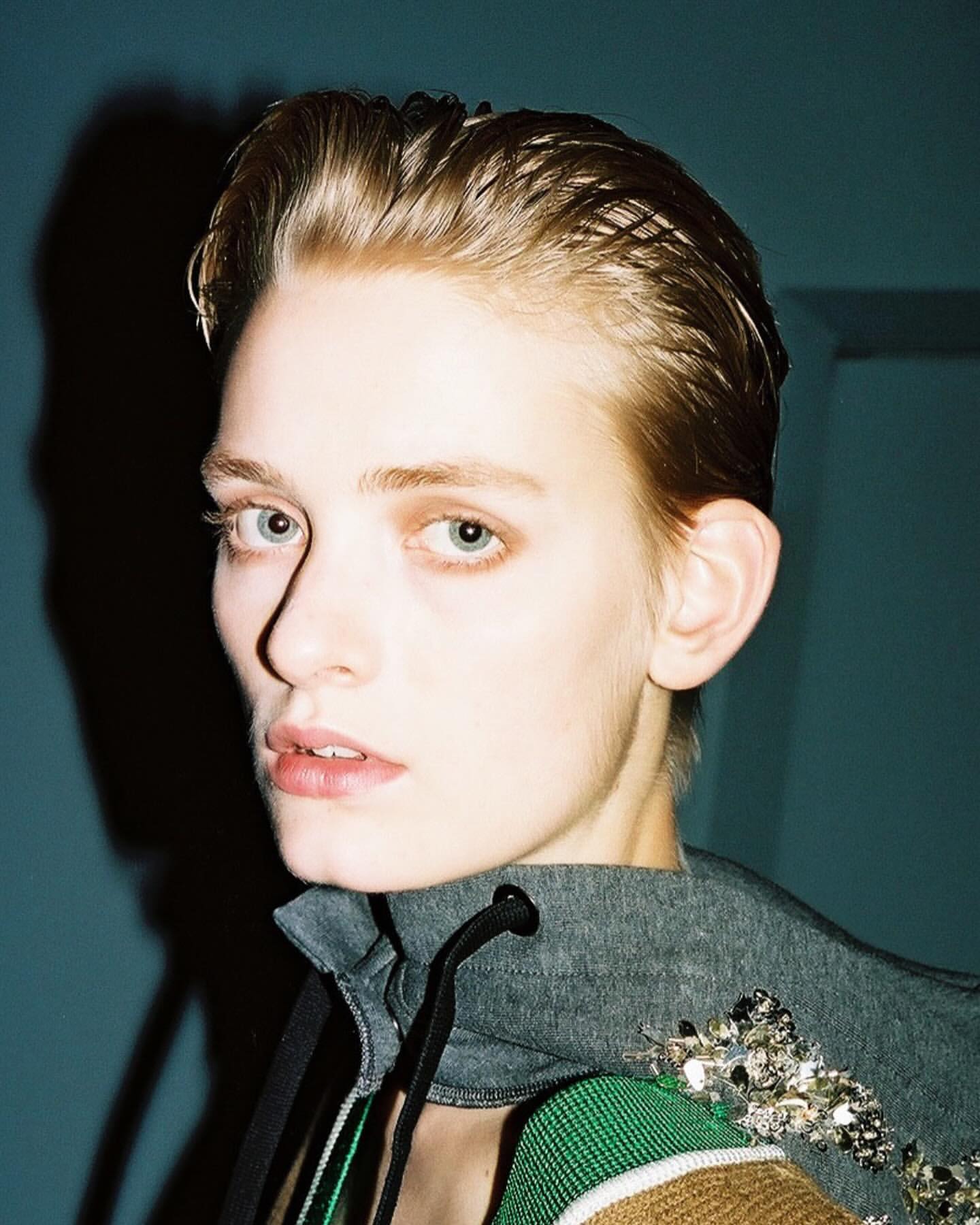 Close-up studio portrait of a young adult female with blonde, slicked-back hair and light skin wearing a gray hoodie with embellished trim.