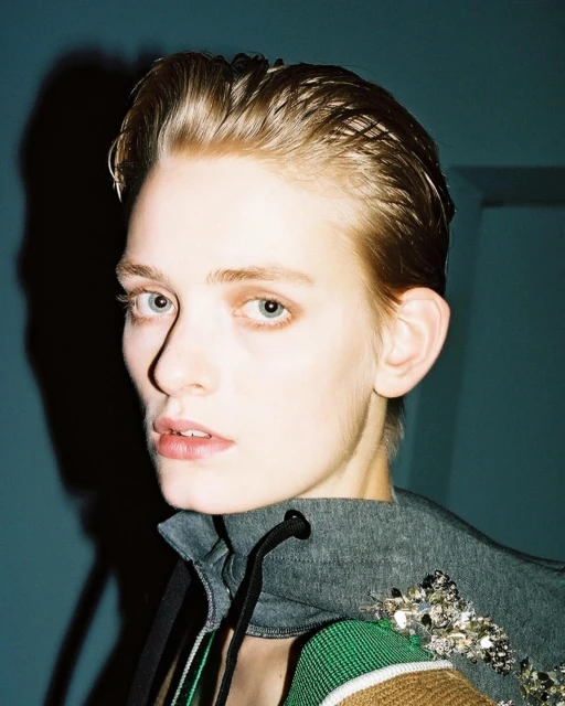 Close-up studio portrait of a young adult female with blonde, slicked-back hair and light skin wearing a gray hoodie with embellished trim.