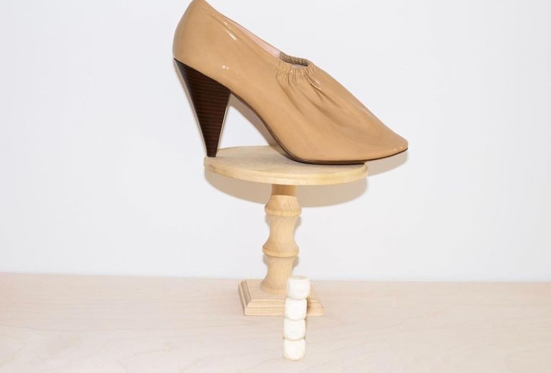Elegant beige patent leather pump shoe with a sculpted heel on a wooden pedestal against a seamless white background, fashion forward.
