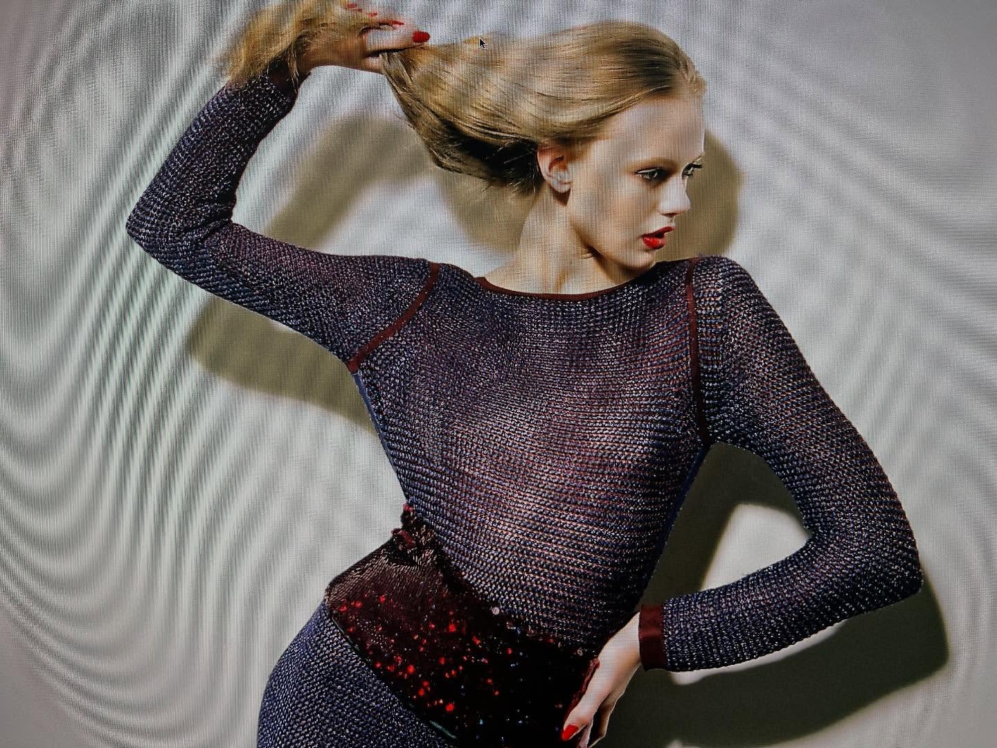 Elegant blonde woman in a textured top posing in front of a mottled wall, highlighted by shadows and soft lighting. Fashion shot.