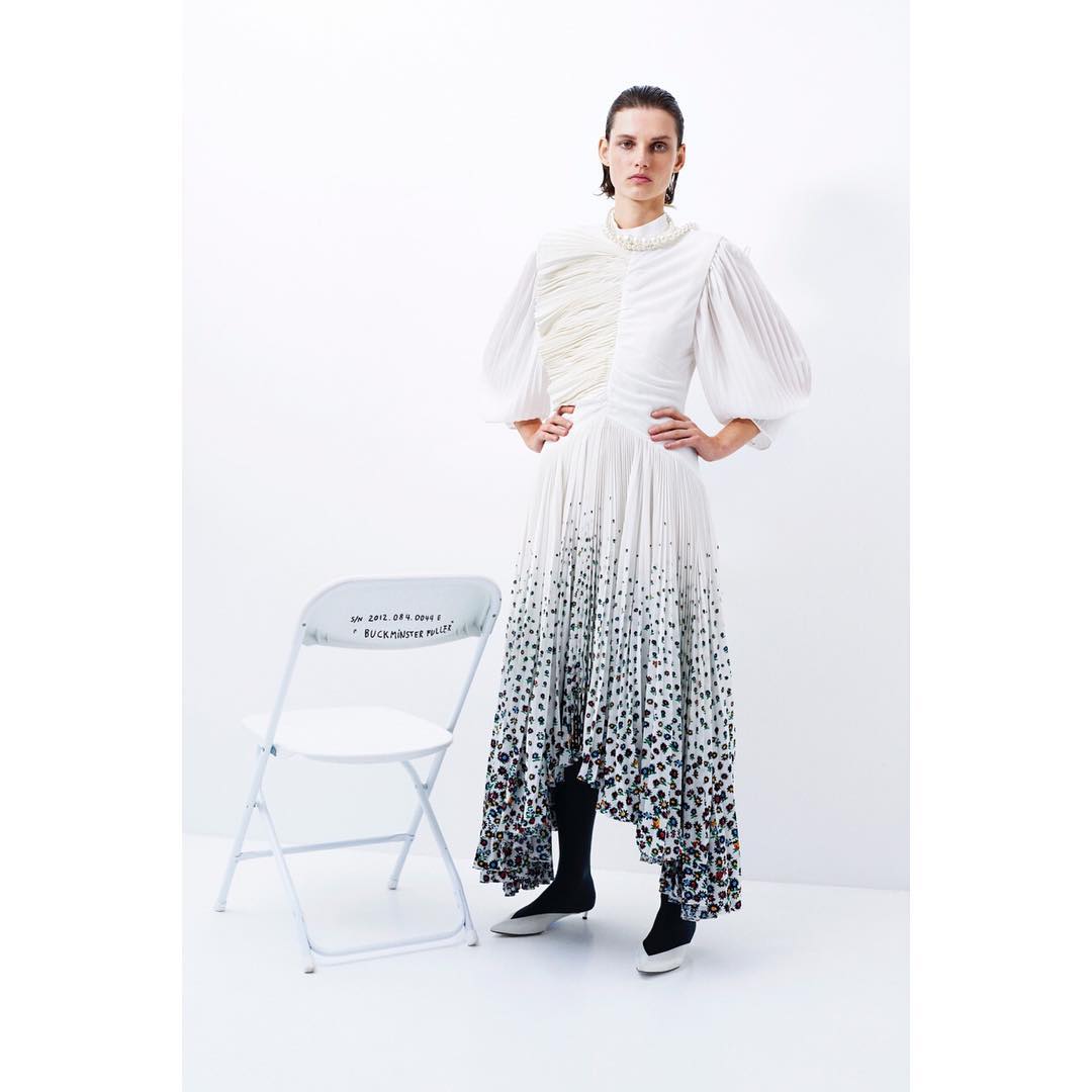 A female model poses indoors in a white patterned dress with puffy sleeves, next to a white folding chair with black text.