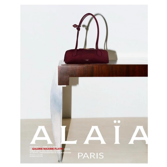 Alaïa burgundy purse is displayed on a dark wood table in a fashion editorial ad against a white backdrop with brand logo.