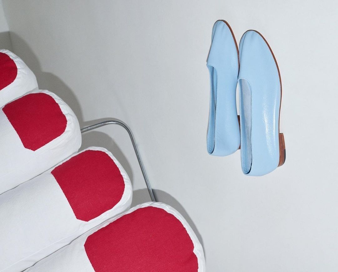 Still life shot of baby blue ballet flats against a white wall with a decorative chair with red and white striped cushions.