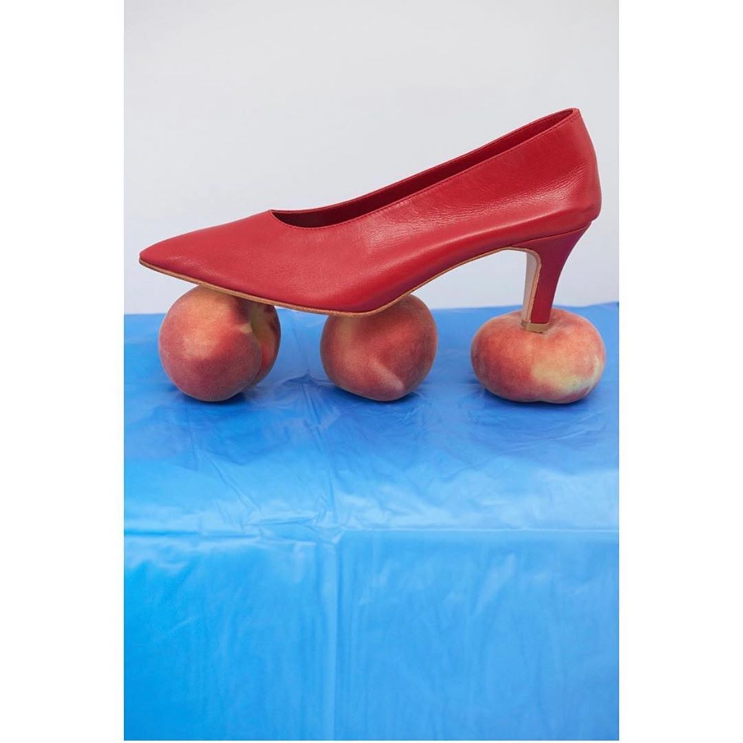 Red high heel shoe perched on three ripe peaches against a blue surface and white background, creating a colorful, conceptual shoe still life.
