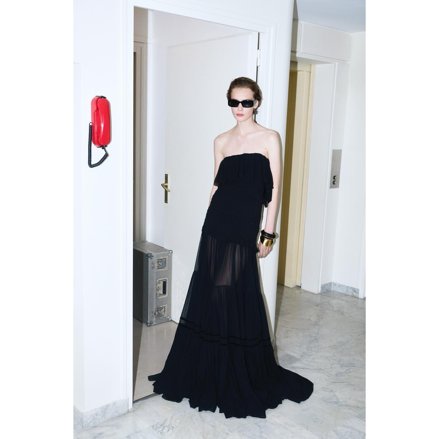A woman in a black strapless dress and sunglasses poses in a white hallway with a red wall phone and silver metal box.
