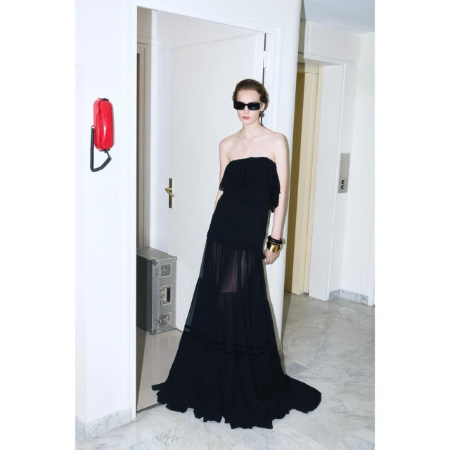 A woman in a black strapless dress and sunglasses poses in a white hallway with a red wall phone and silver metal box.