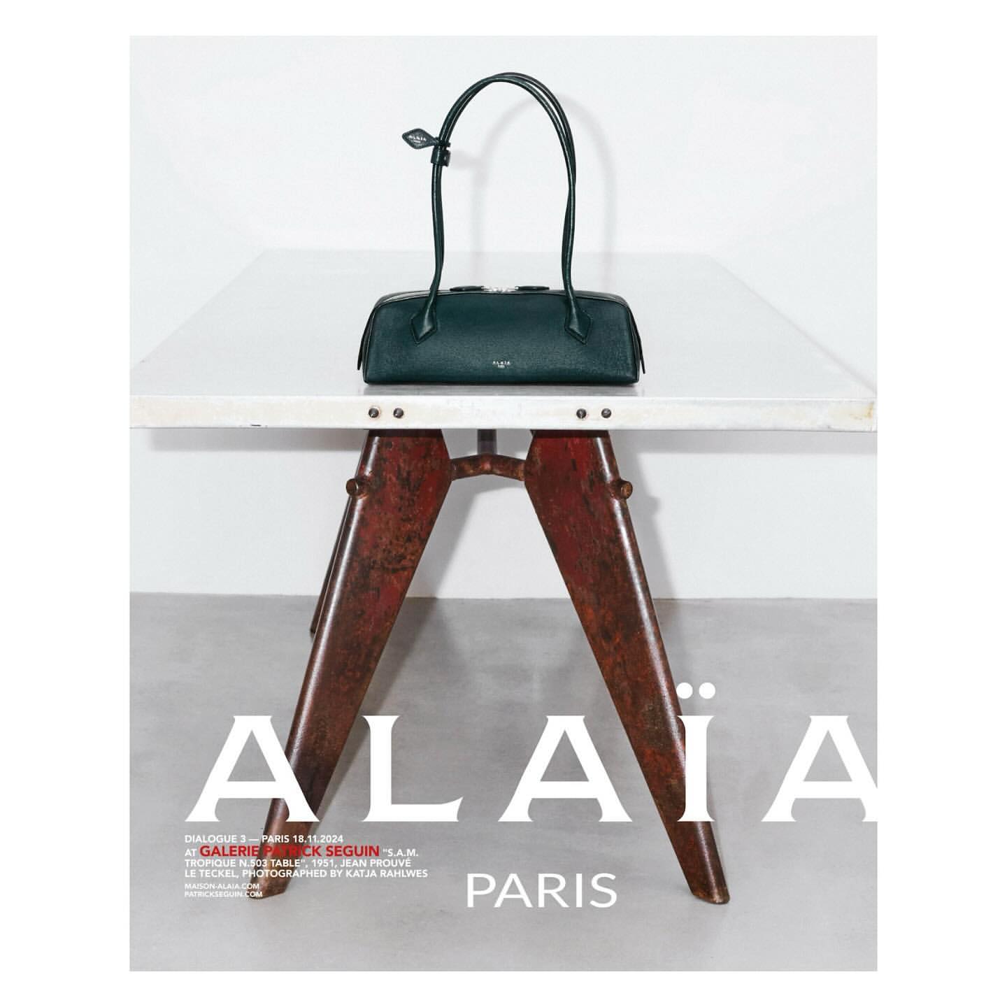 Alaïa handbag in dark green leather with thin top handles on a white table with rusted metal base against a white wall.