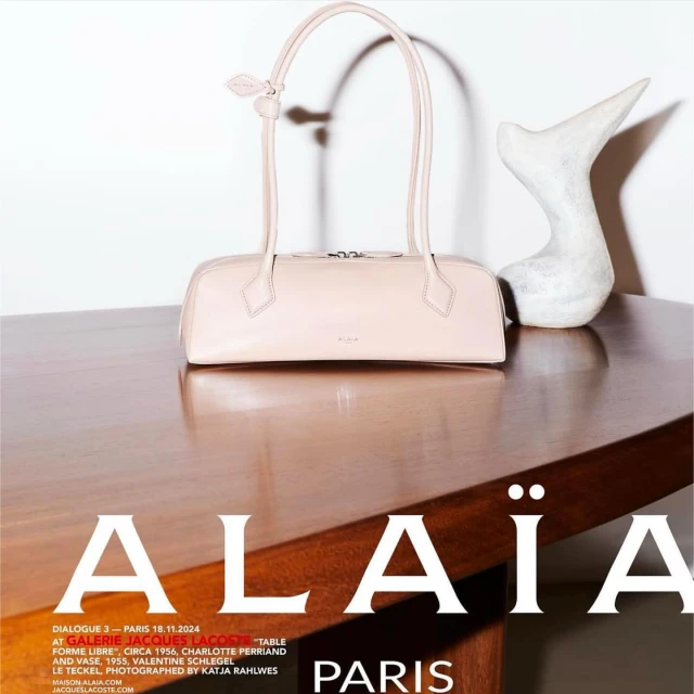 Elegant cream-colored leather handbag with dual top handles on a polished wood table, paired with a modern white sculpture.