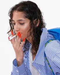 Close-up portrait of a woman with wet dark hair, bright red long nails, smoking a cigarette, wearing a blue and white striped shirt.