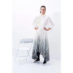 Model in white pleated dress with dark floral hem standing next to a white folding chair against a white background
