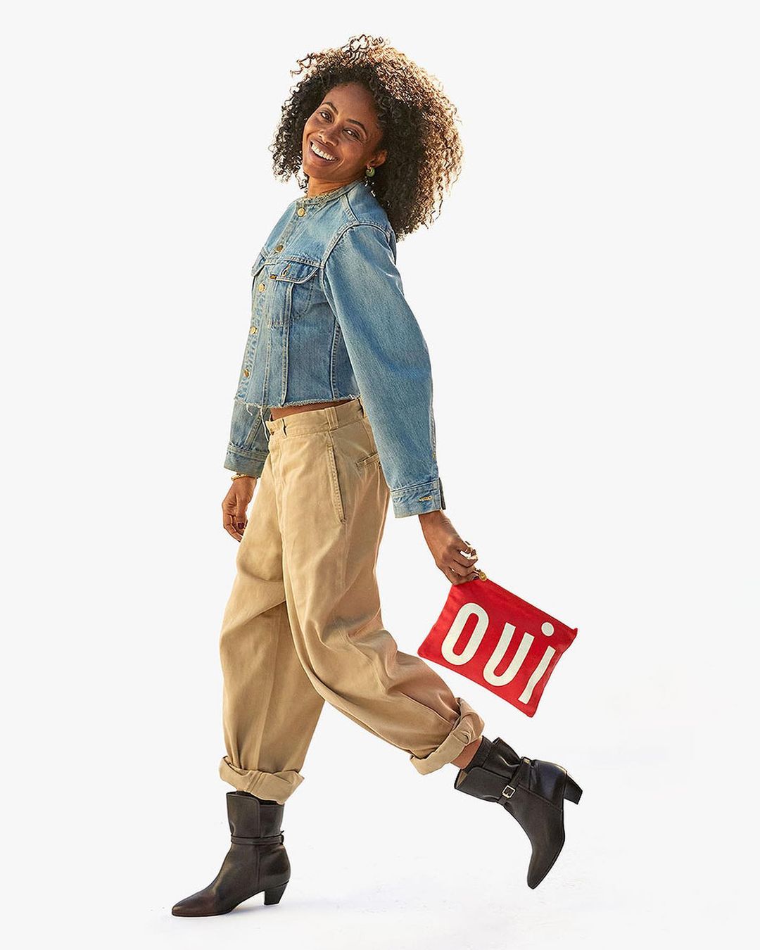 A smiling African American woman in a denim jacket and tan pants walks with a red 'oui' pouch against a white background.