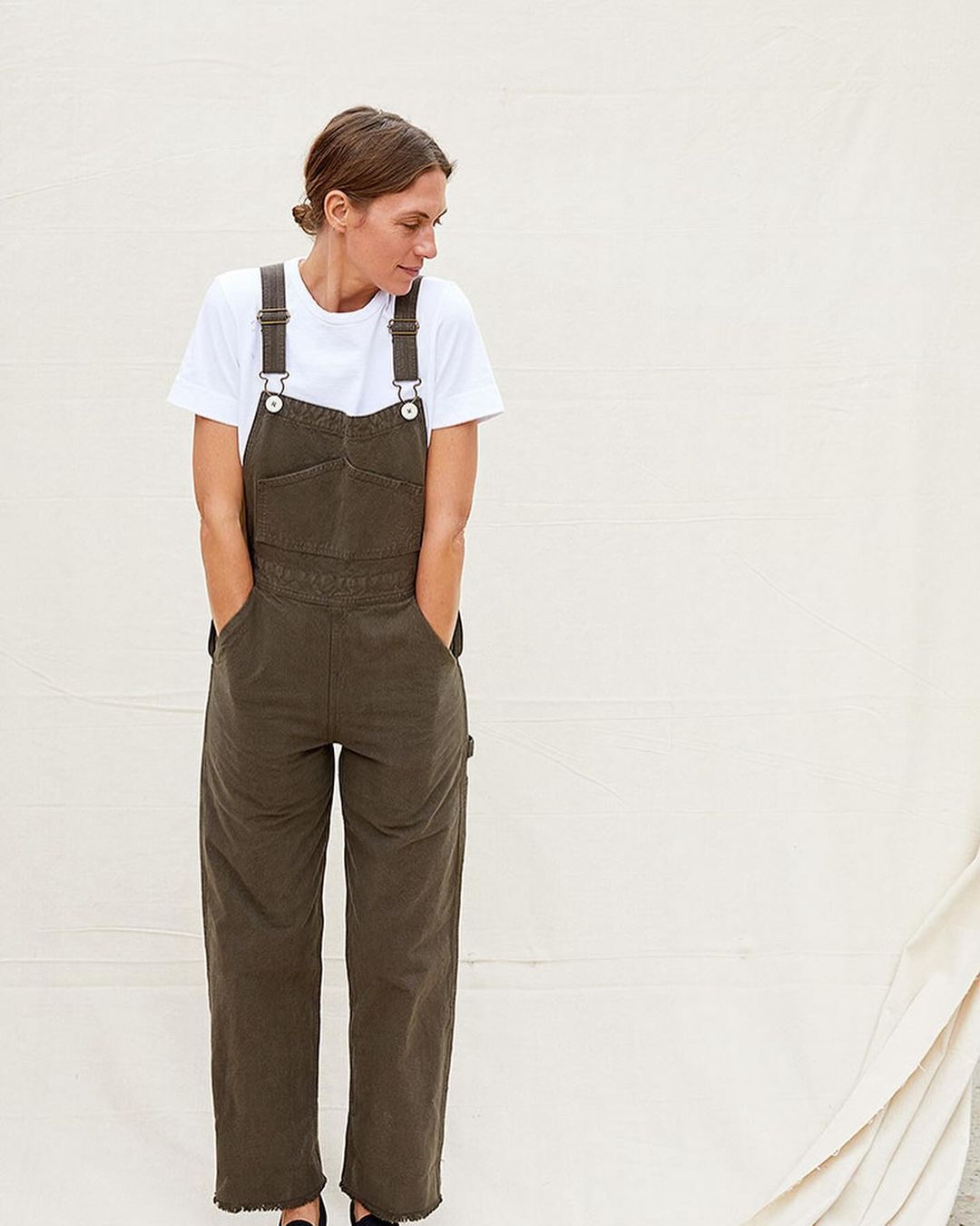 A fair-skinned woman with her hands in her pockets poses in olive-green denim overalls and a white t-shirt against a cream-colored background.