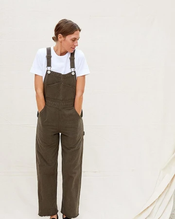 A fair-skinned woman with her hands in her pockets poses in olive-green denim overalls and a white t-shirt against a cream-colored background.