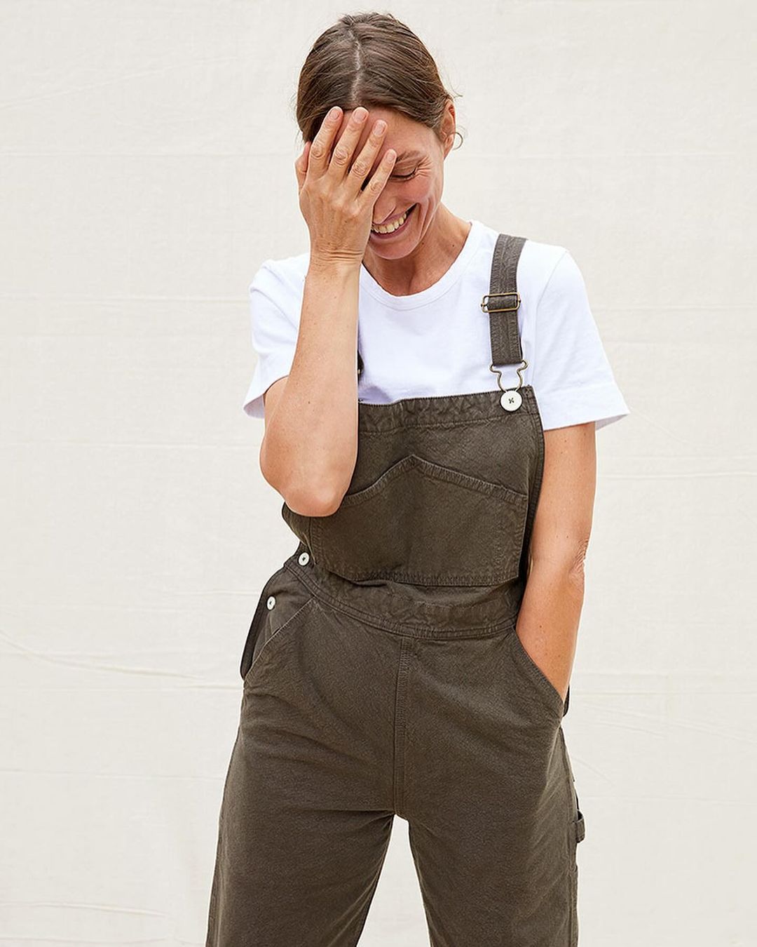 Smiling woman wearing dark green overalls over a white t-shirt with her hand covering her eyes against a white wall.