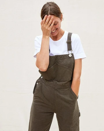 Smiling woman wearing dark green overalls over a white t-shirt with her hand covering her eyes against a white wall.