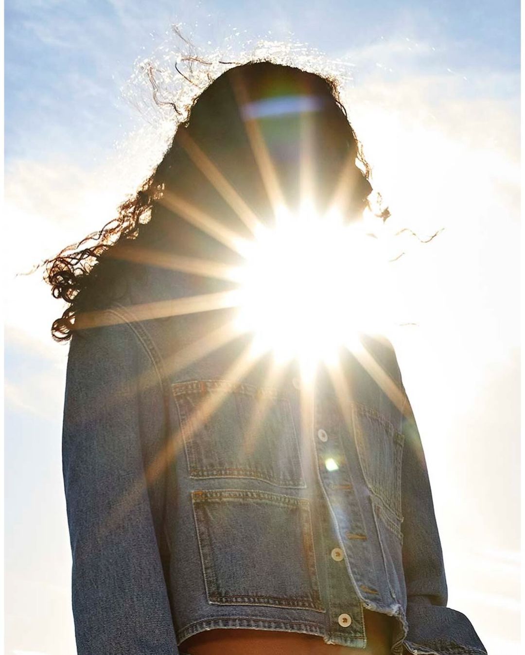 Woman backlit by the sun wearing a denim jacket against a clear sky. The bright light creates a lens flare effect.