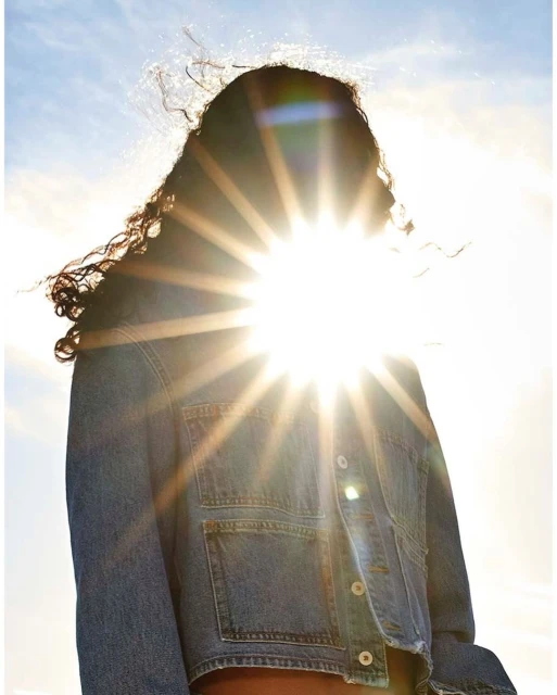 Woman backlit by the sun wearing a denim jacket against a clear sky. The bright light creates a lens flare effect.