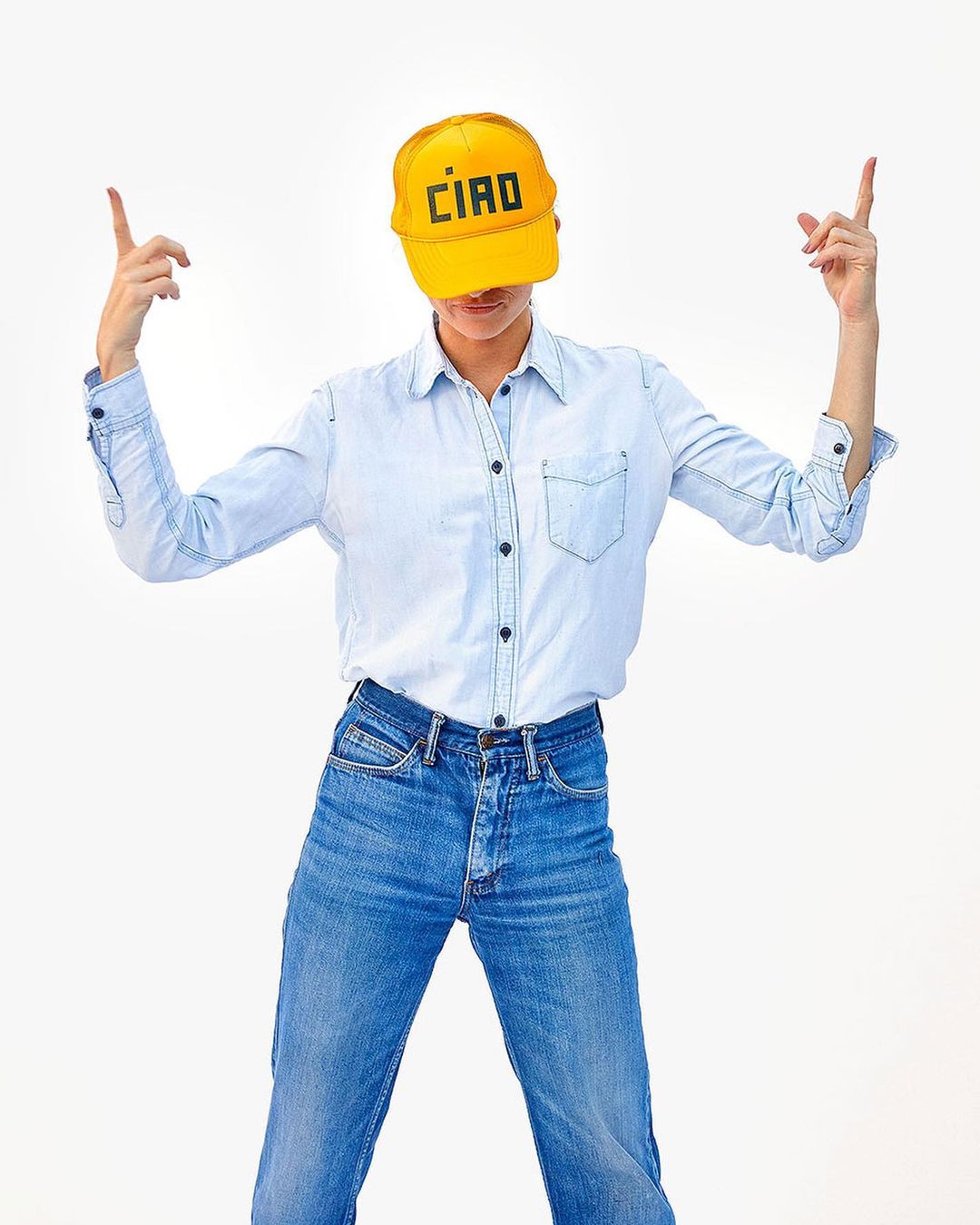 Adult woman with a yellow cap, denim shirt, and jeans stands with both arms raised on a solid white background in a studio shot.