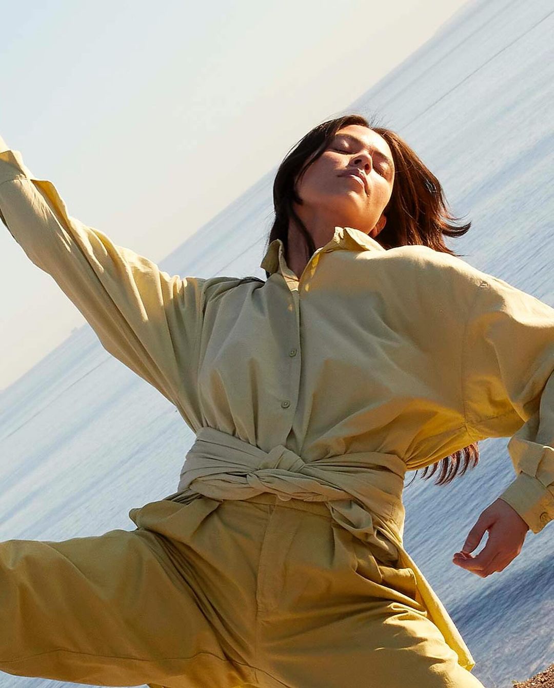 A woman in a yellow blouse and pants stands with her arms outstretched against a background of the sea and sky in an outdoor setting.