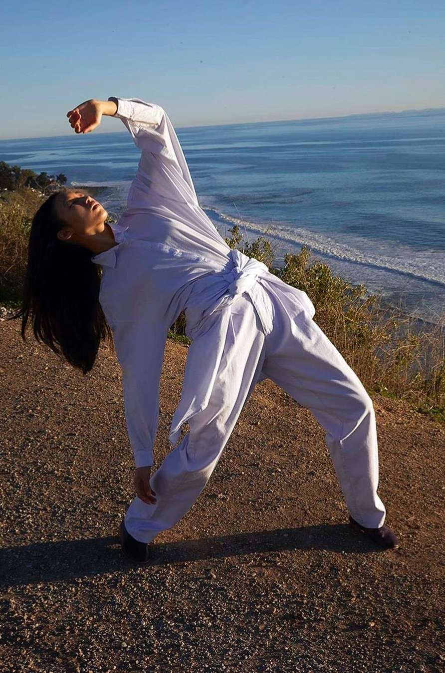Woman in white shirt and pants posing dramatically on a cliff overlooking the ocean with clear blue water