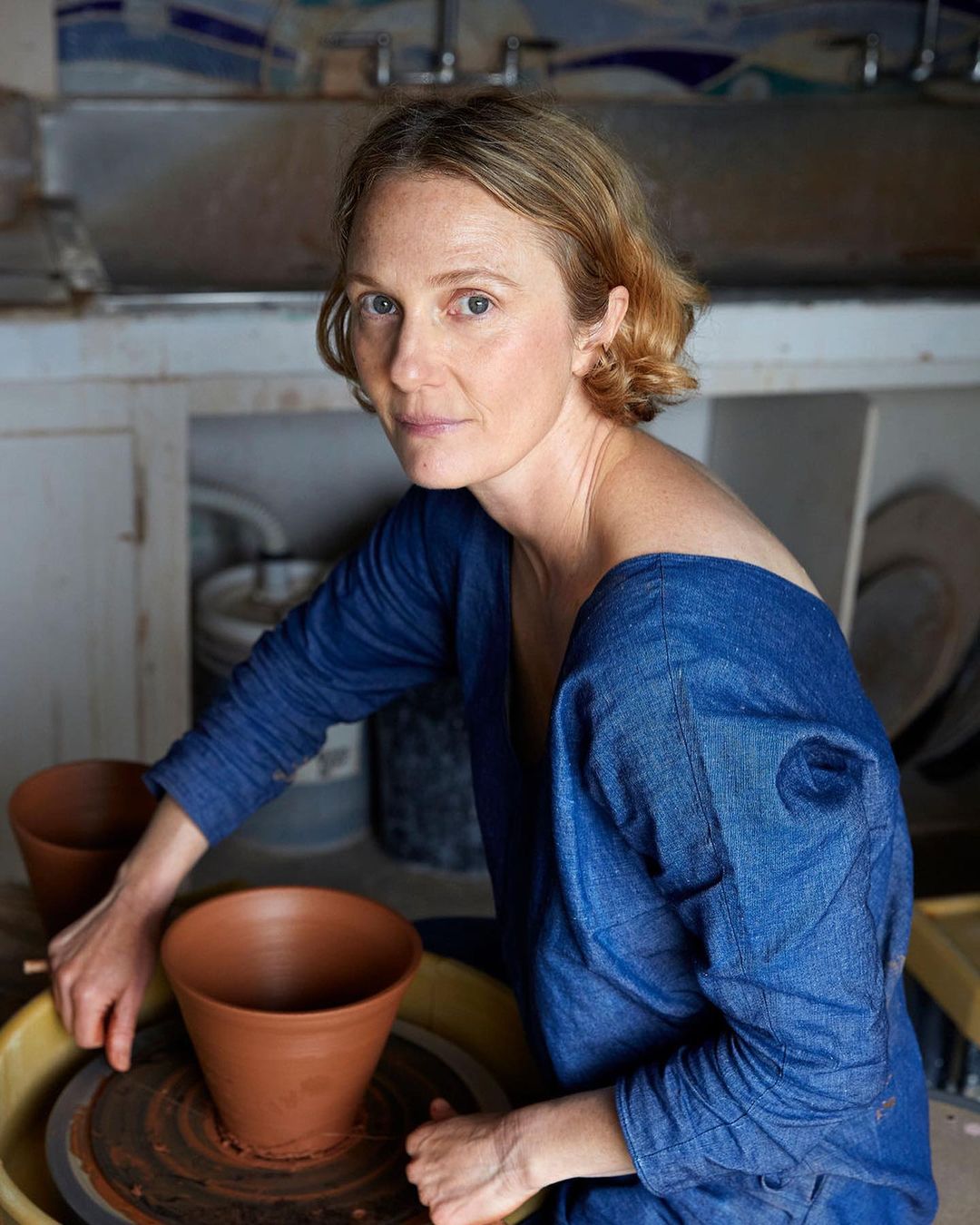 Woman creating pottery on a pottery wheel, wearing a blue shirt. The pottery is reddish brown and the setting is indoors.