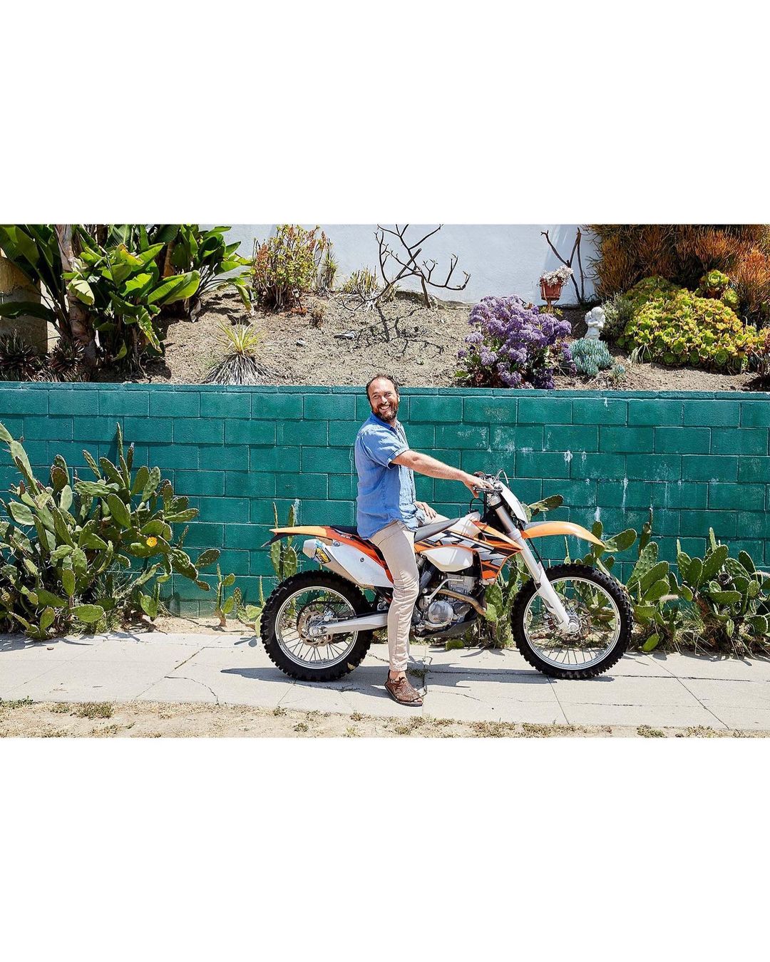 An adult man riding a white and orange dirt bike on a sidewalk, with a green cinderblock wall and garden in the background on a sunny day.