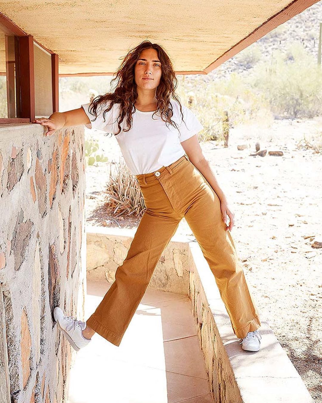 Adult female in a white shirt and tan pants leans against a stone wall, posing outdoors in a desert landscape with hills in the background.
