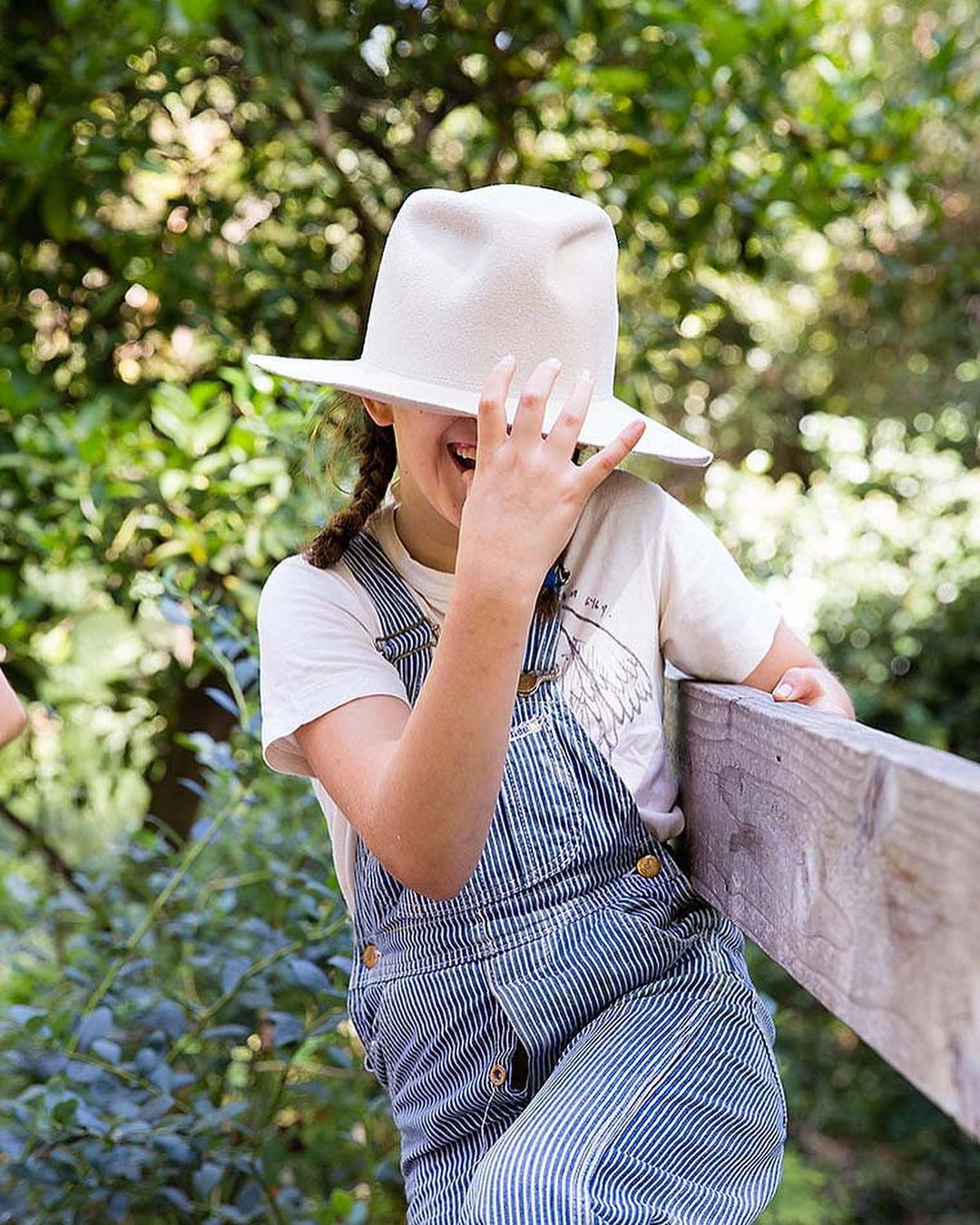 A child with braids wearing overalls and a white shirt is partially obscured by a cream-colored cowboy hat and is smiling widely.
