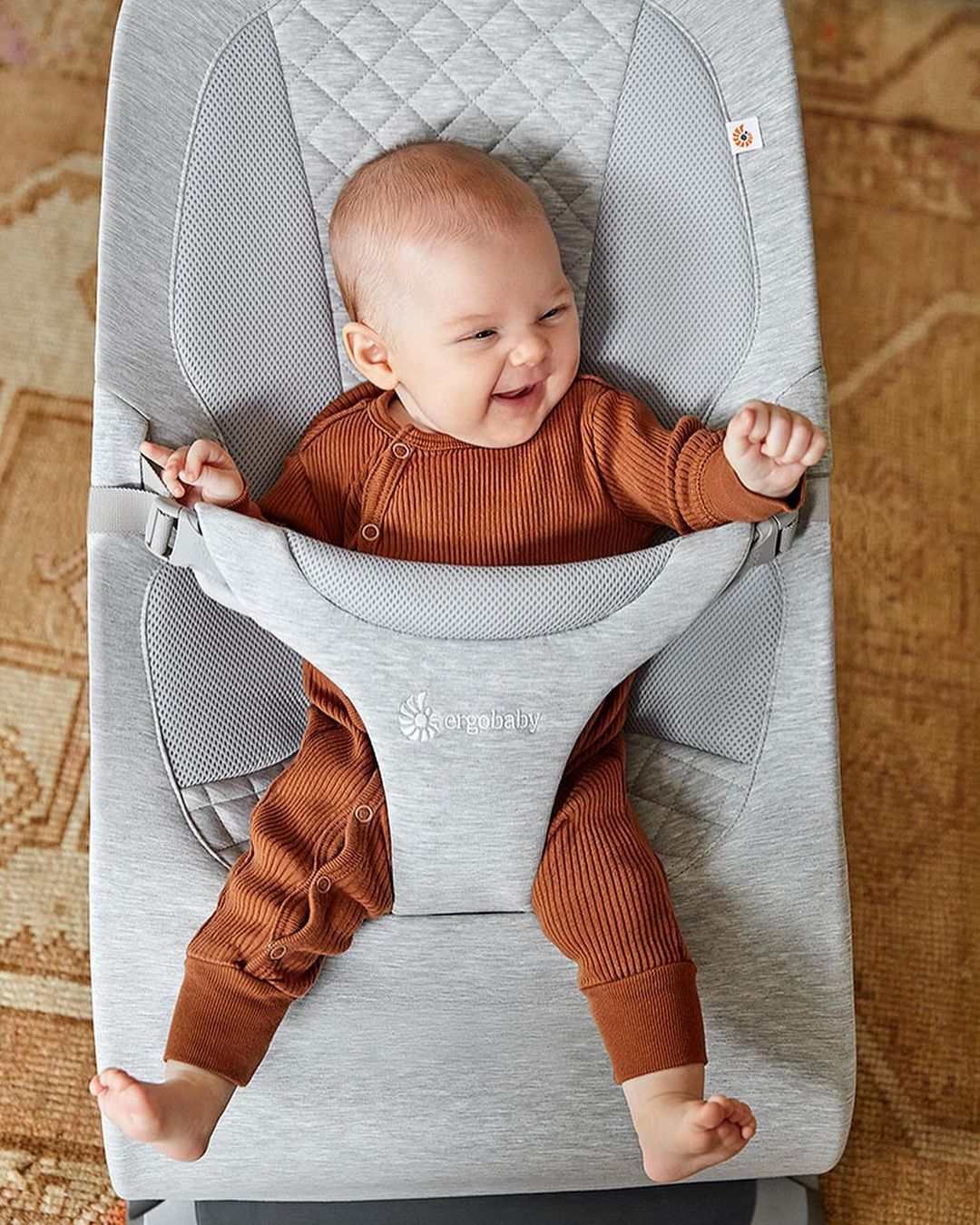 Infant in a brown ribbed onesie smiles while sitting in a gray Ergobaby bouncer on a patterned rug in a home setting.