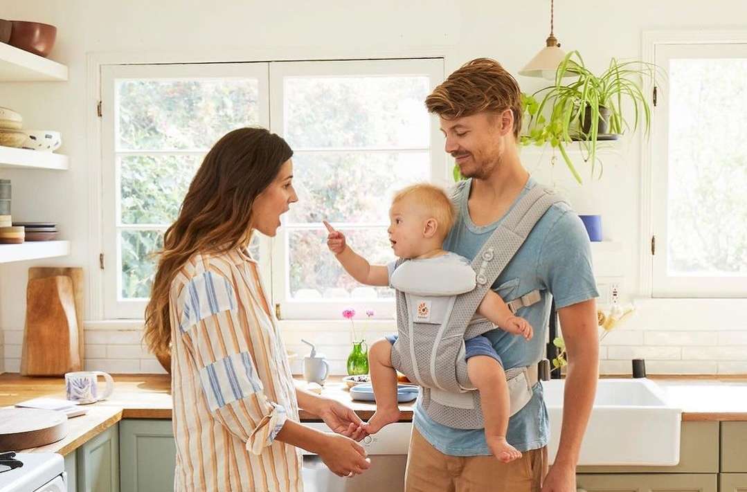 Family of three in a bright kitchen: A father holding a baby in a carrier, and a mother interacting with them in a home interior.