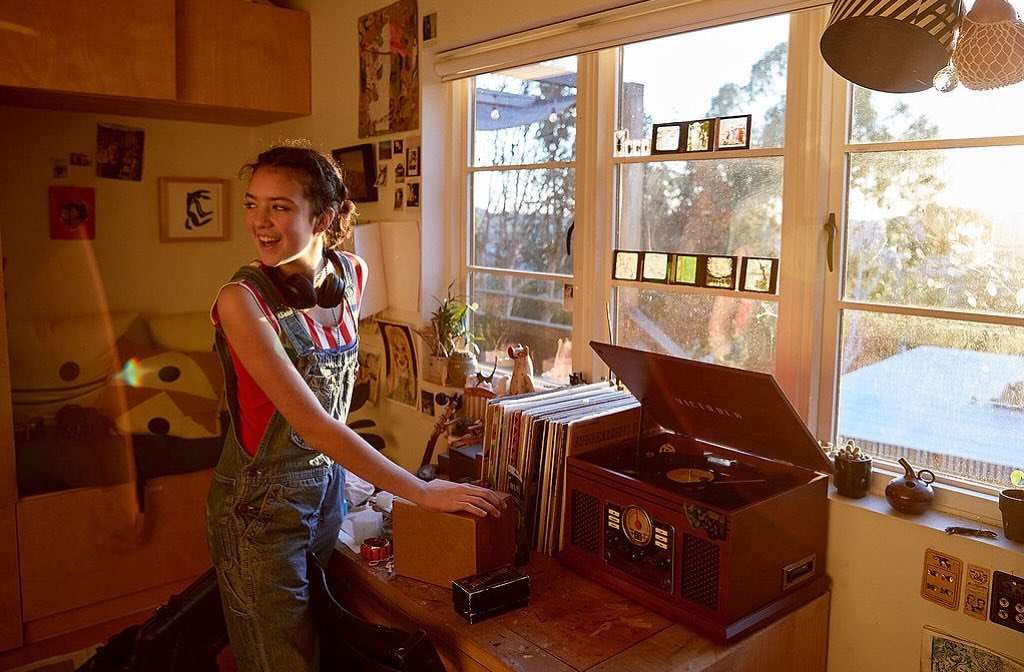 Smiling woman in overalls stands near a record player in a sunlit room with various objects and a window view of trees and a blue sky.