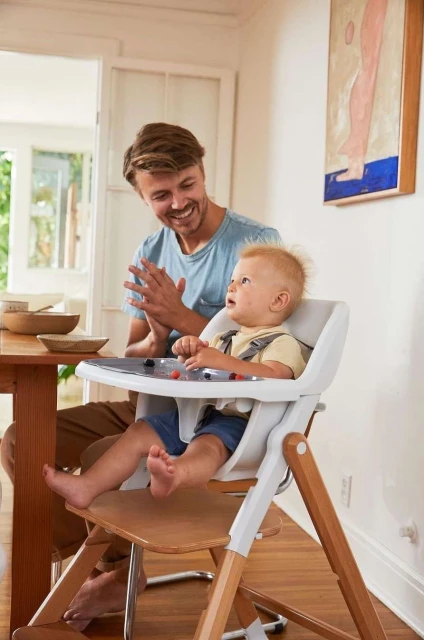 A fair-skinned baby with blonde hair sits in a high chair at a table indoors, with a man smiling in the background. 