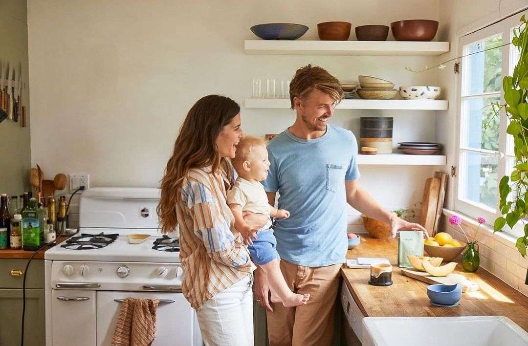 Family of three, mother holding baby, father, standing in a light-filled kitchen next to a counter with fruit and plants. 