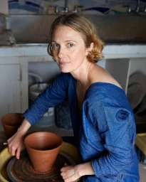 A woman in a blue denim top shaping wet clay on a pottery wheel in a studio workshop.
