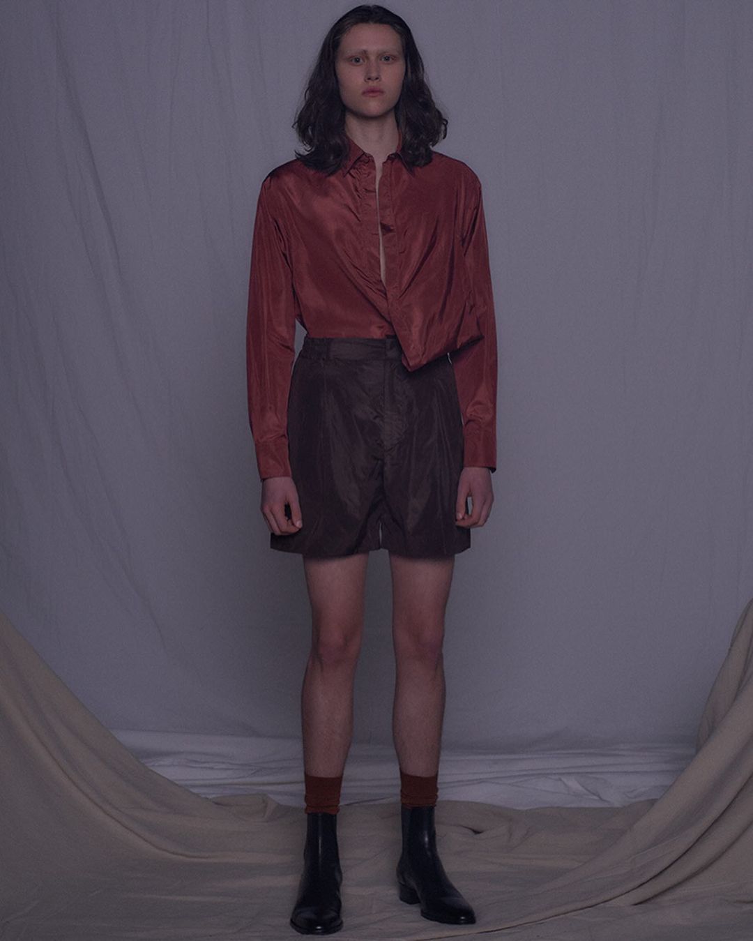 Full-length studio shot of a slender person in a red button-down shirt, brown shorts, brown socks, and black boots against a draped backdrop.