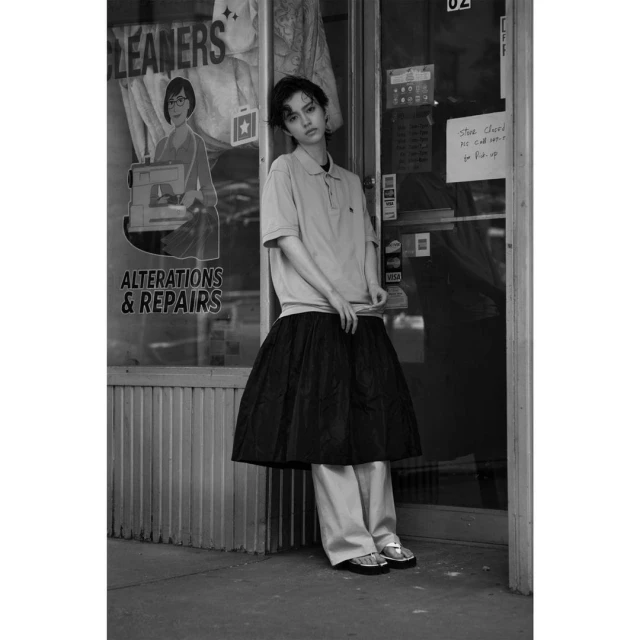 Young woman stands in front of a dry cleaner, wearing a polo shirt, full black skirt, and light-colored pants and silver sandals.