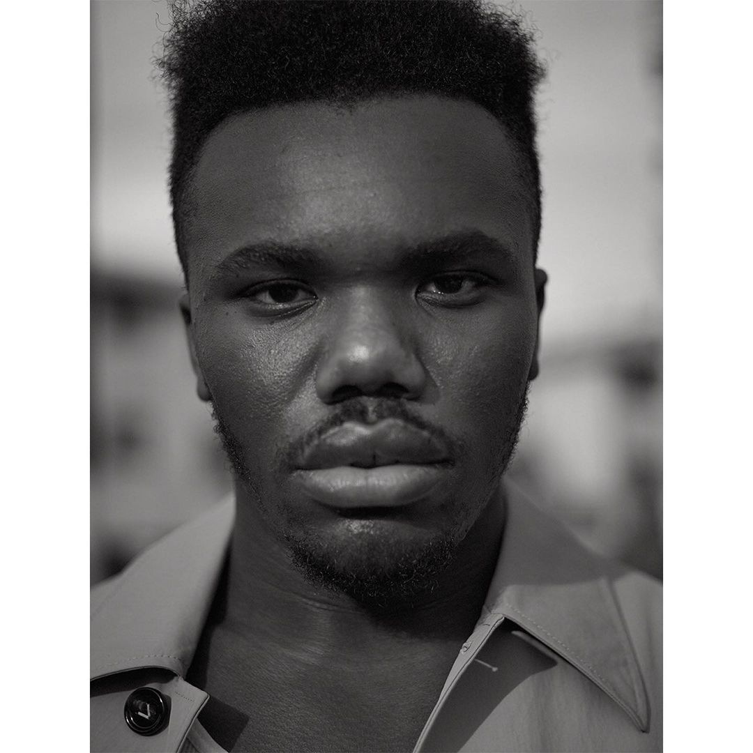 Portrait of a young adult Black man in a collared shirt, looking directly at the camera in a close-up, monochrome studio shot.