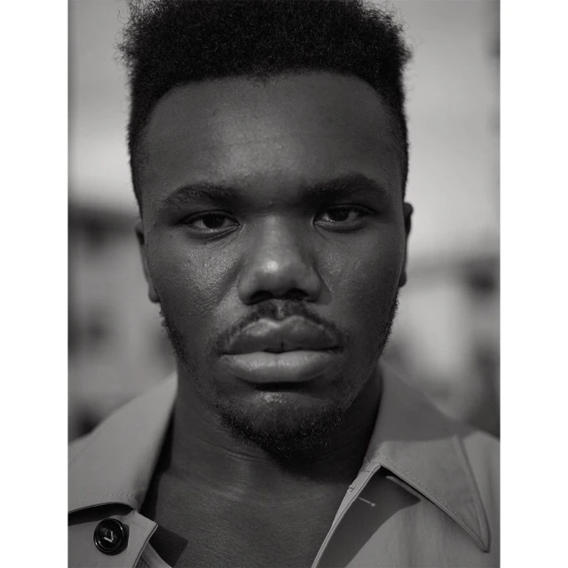 Portrait of a young adult Black man in a collared shirt, looking directly at the camera in a close-up, monochrome studio shot.