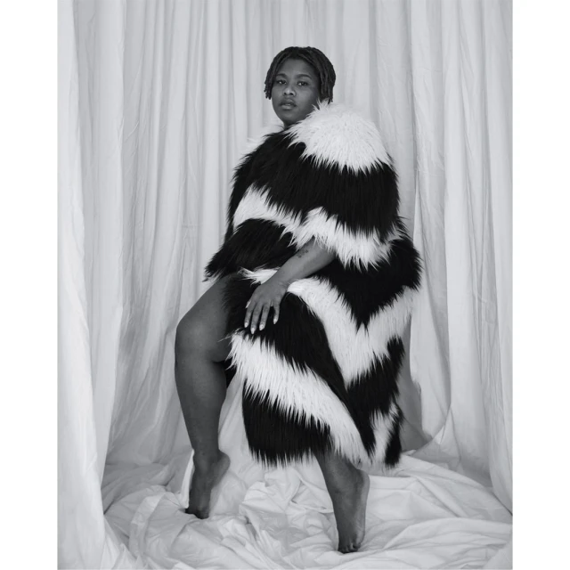 Monochromatic portrait of a woman draped in a striking black and white chevron-patterned faux fur coat, posing against a draped fabric backdrop.