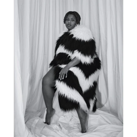 Monochromatic portrait of a woman draped in a striking black and white chevron-patterned faux fur coat, posing against a draped fabric backdrop.