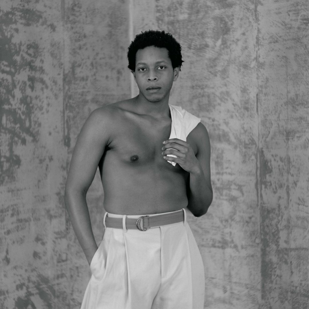 Portrait of a shirtless Black man wearing light-colored trousers against a textured concrete wall by photographer Katsu Naito.