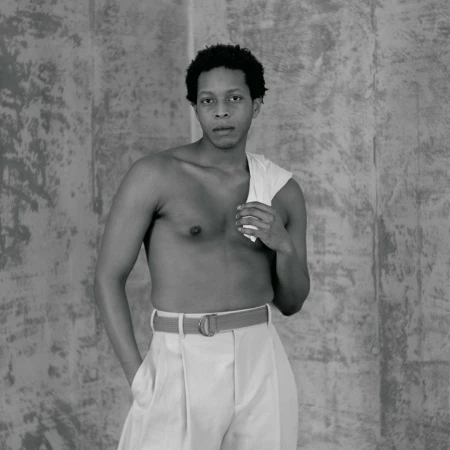 Portrait of a shirtless Black man wearing light-colored trousers against a textured concrete wall by photographer Katsu Naito.